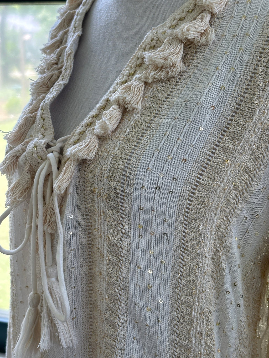 Close-up of Buby.S tassel tie V-neckline with gold sequin detail and ladder stitch weave on ivory tunic