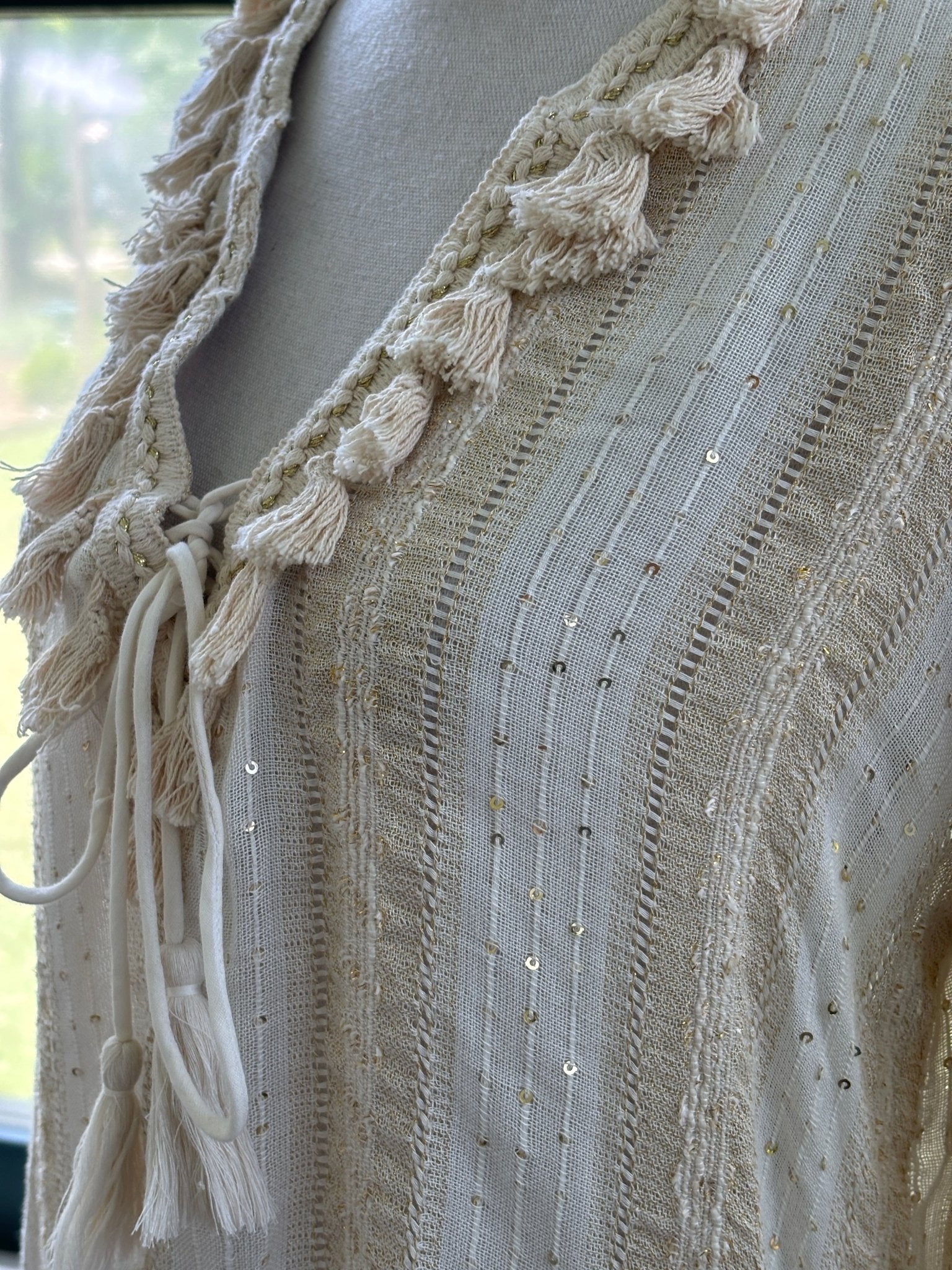 Close-up of Buby.S tassel tie V-neckline with gold sequin detail and ladder stitch weave on ivory tunic