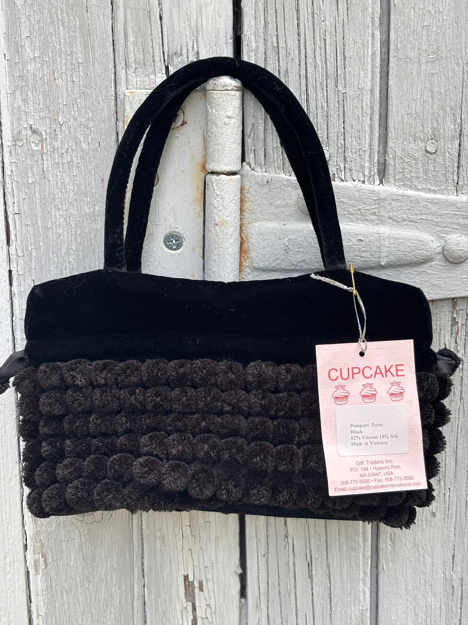 Cupcake International black pompom purse hanging on weathered fence, dual velvet top handles and dense pompom body, NWT
