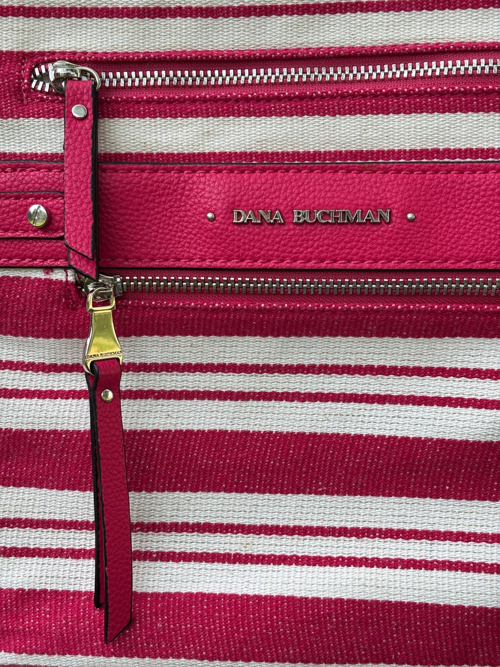 Close-up of Dana Buchman embossed logo plate, dual zip pockets with gold and silver hardware, and leather zipper pull on red stripe canvas crossbody