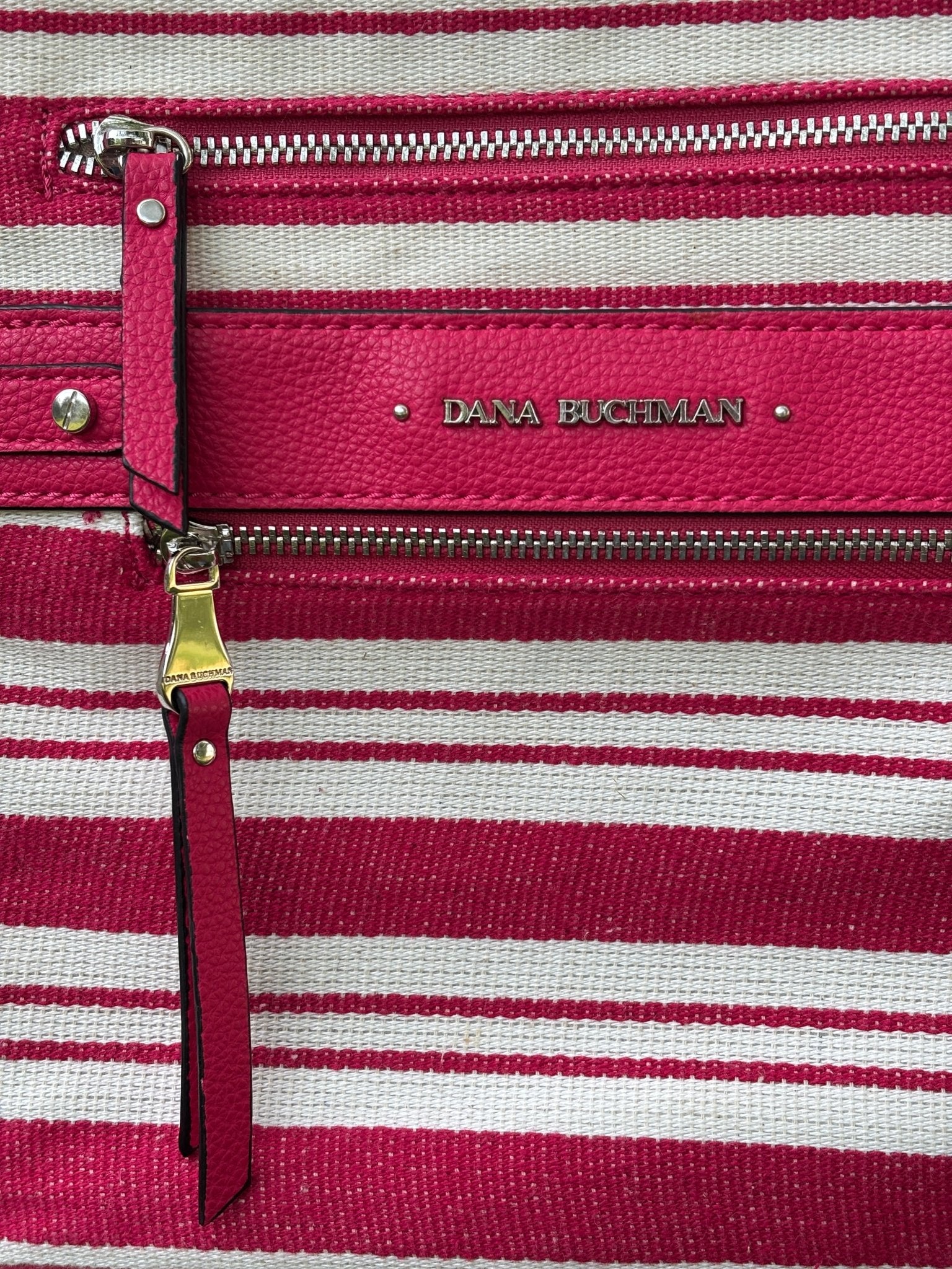 Close-up of Dana Buchman embossed logo plate, dual zip pockets with gold and silver hardware, and leather zipper pull on red stripe canvas crossbody