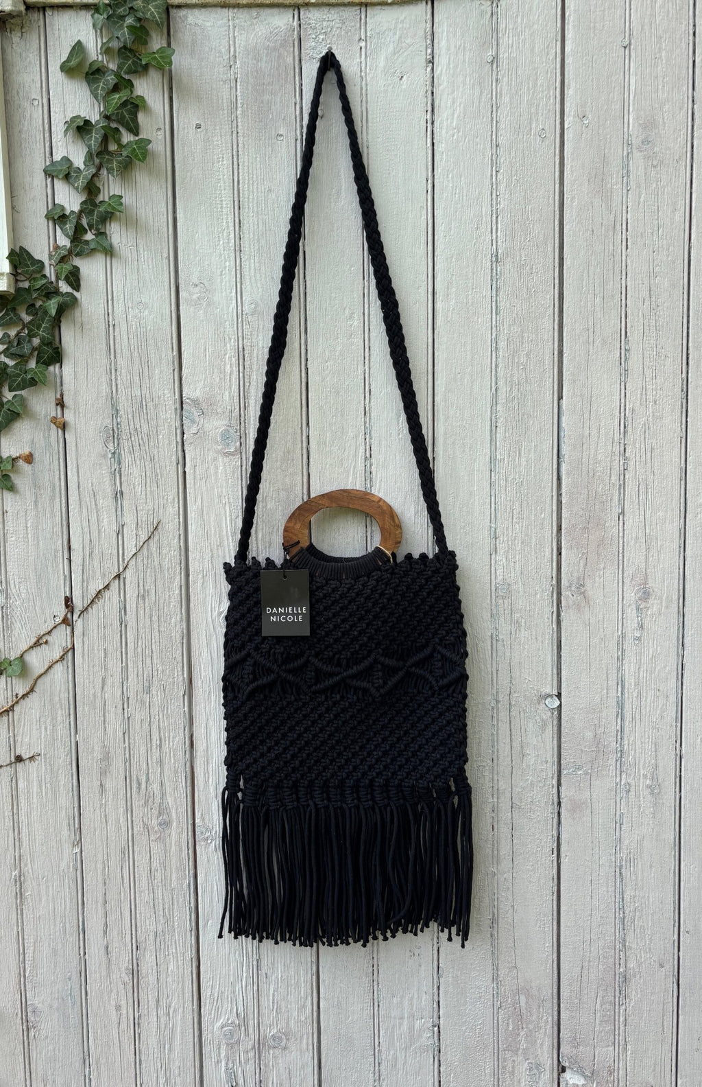 Danielle Nicole black macramé fringe tote bag hanging on weathered fence, round wood handle and braided crossbody strap, NWT