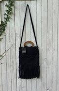 Danielle Nicole black macramé fringe tote bag hanging on weathered fence, round wood handle and braided crossbody strap, NWT