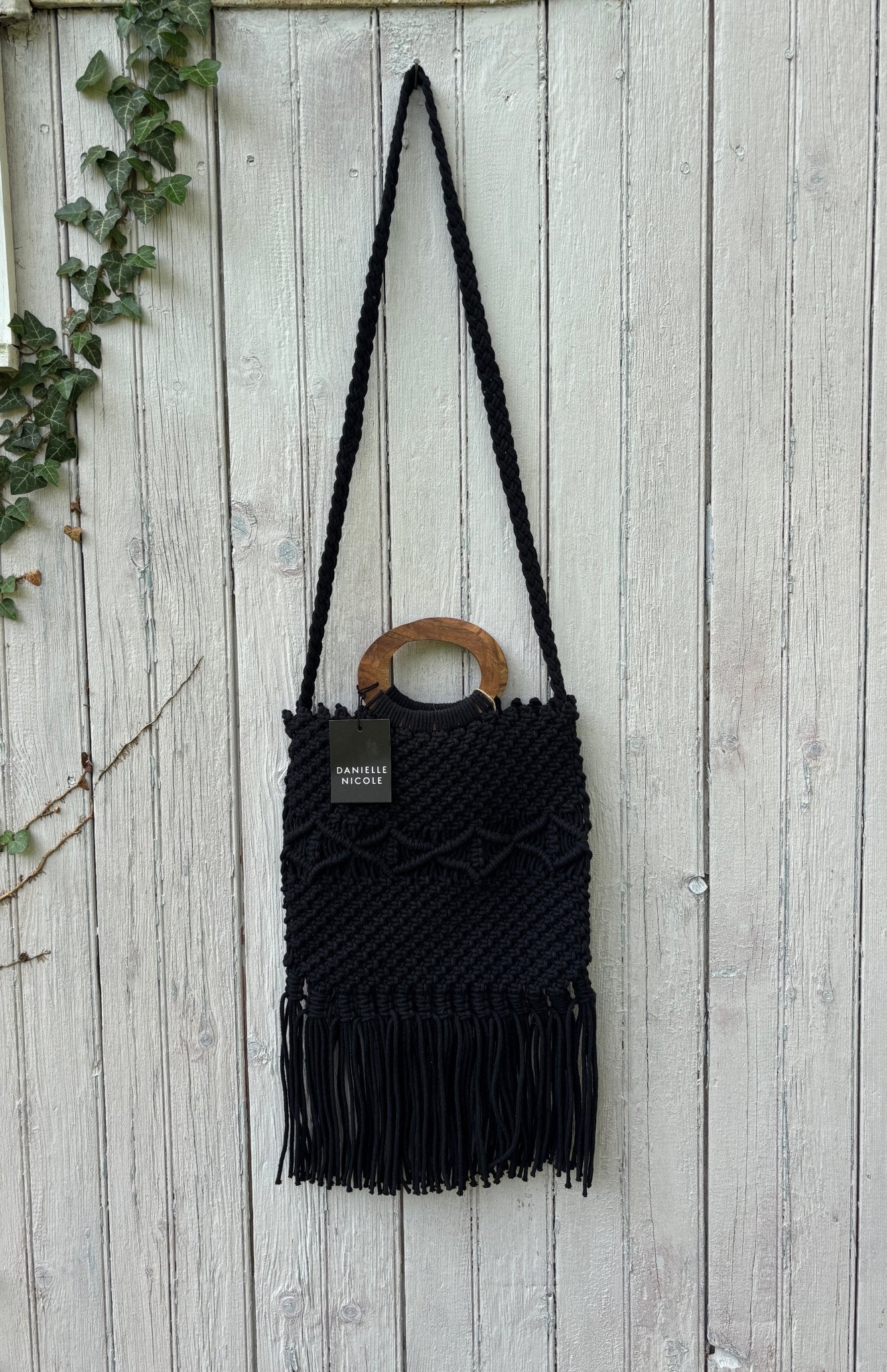 Danielle Nicole black macramé fringe tote bag hanging on weathered fence, round wood handle and braided crossbody strap, NWT